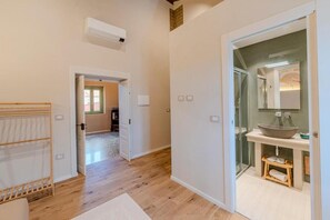 Quarto de hotel com piso de madeira, ar condicionado branco, box de vidro e prateleira de madeira com toalhas.