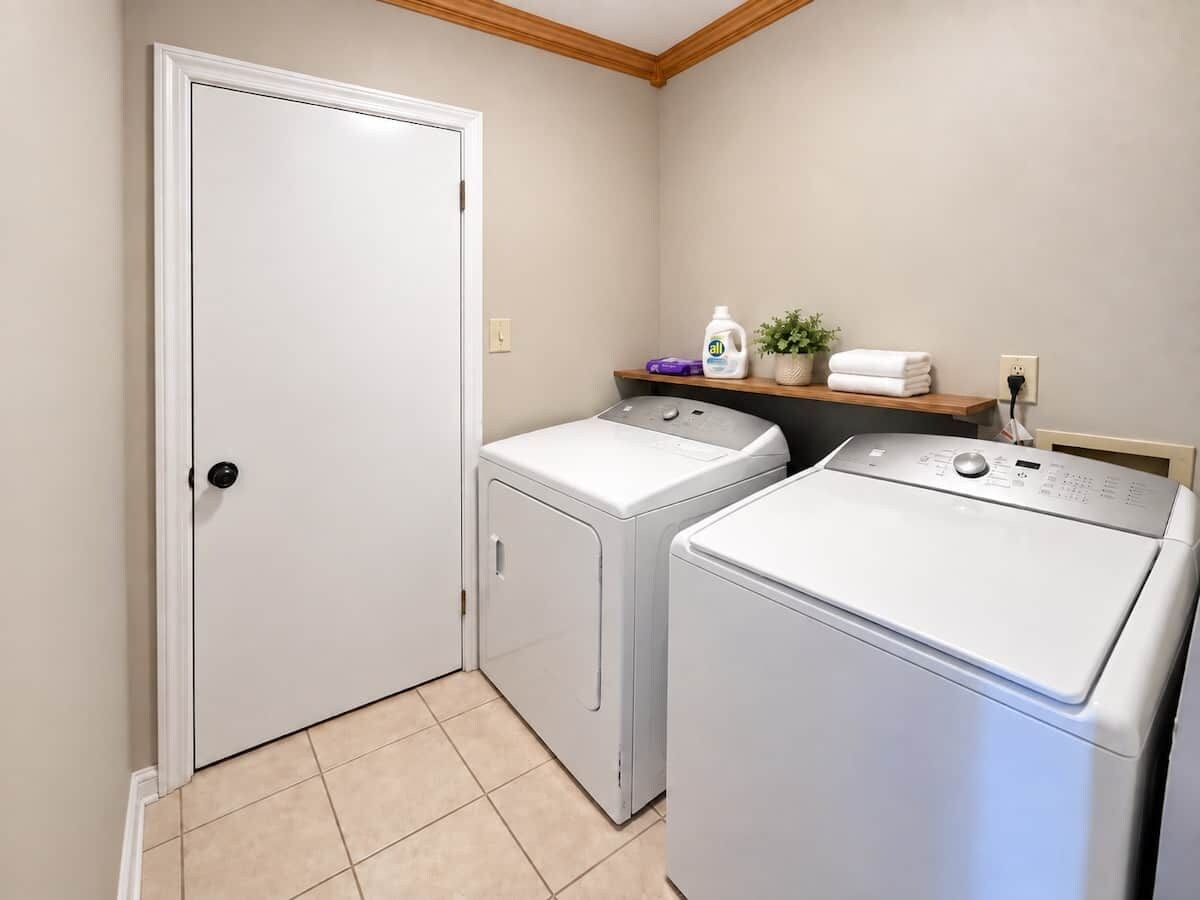 A laundry room with a door, two washing machines, a shelf with a plant, and a bottle of detergent.
