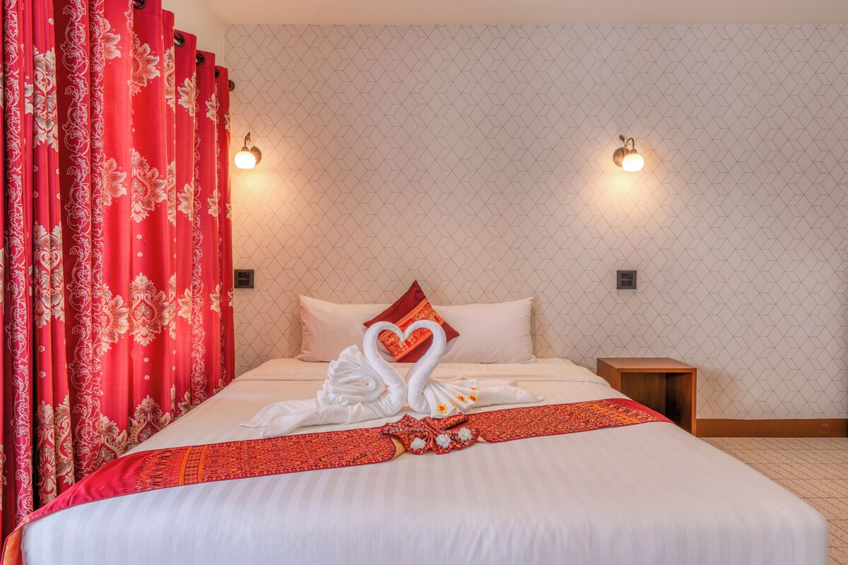 Una habitación de hotel con una cama, cortinas rojas, una toalla dispuesta en forma de corazón y dos luces fijadas a la pared.