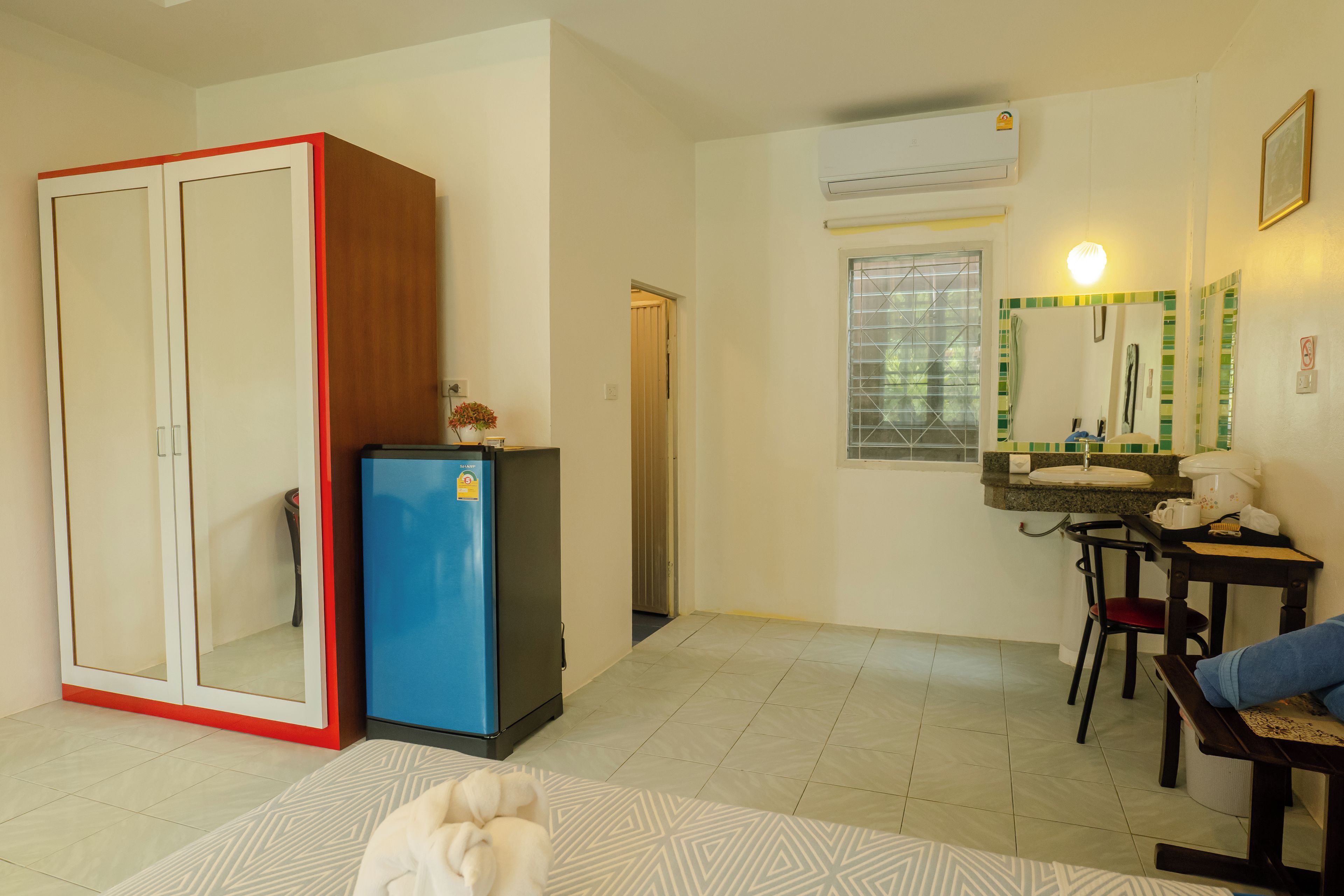A compact living space with a kitchenette, a wardrobe, a small fridge, a dining area with a table and chairs, and a bathroom with a mirror and window.