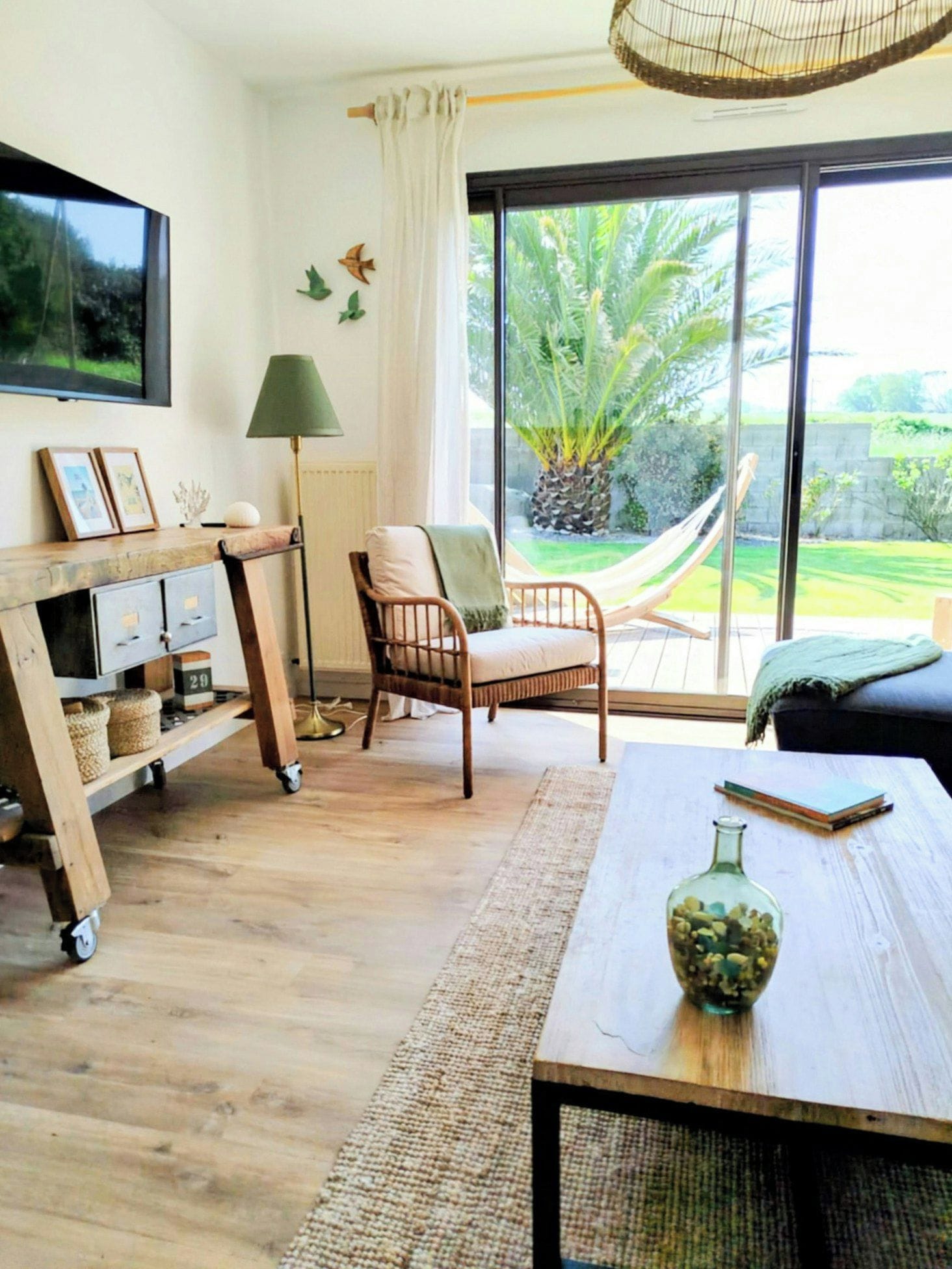 A room with a large window, a hammock, a chair, a table with a vase, and a lamp.