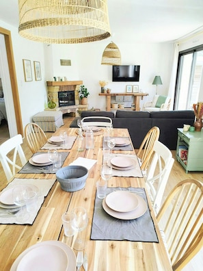 Een eetgedeelte met een houten tafel gedekt voor vier personen, omringd door witte stoelen, met een televisie en een bank op de achtergrond.