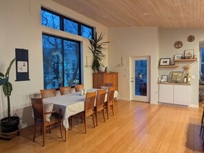 Un comedor con mesa y sillas de madera, ventanas amplias y vista a árboles.