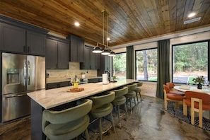 A modern kitchen with a central island, bar stools, and a dining area with a table and chairs. - Broke `n Boujee · New Listing! Heated Pool, Sauna, Arcade & Mini-Golf for 20 (Broken Bow)