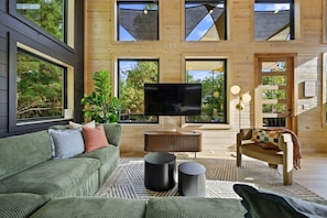 A modern living room with a large flat-screen TV, a green sofa, a patterned rug, and a wooden floor. - Broke `n Boujee · New Listing! Heated Pool, Sauna, Arcade & Mini-Golf for 20 (Broken Bow)
