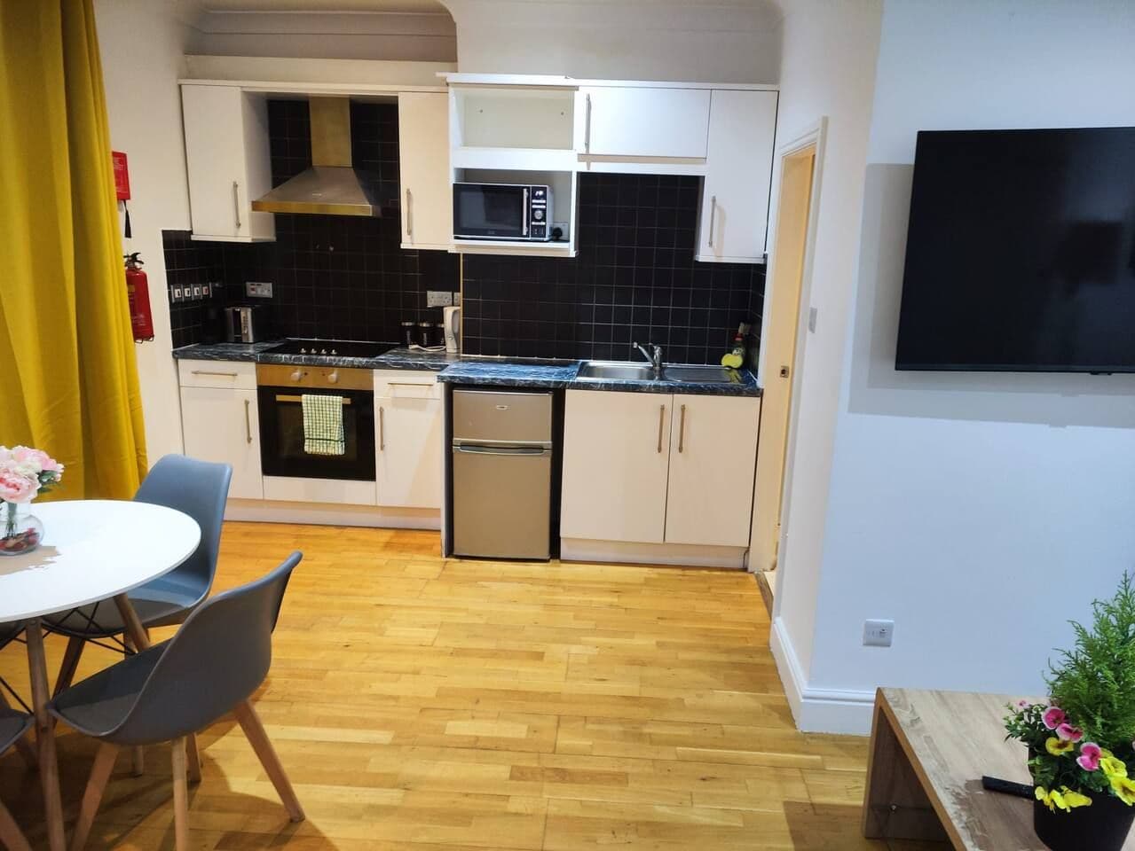 A compact kitchen with a microwave, stove, and sink. There is a dining table with chairs and a potted plant.
