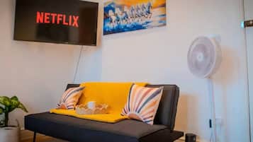A living room with a black sofa, a yellow throw blanket, a painting, a TV displaying 'NETFLIX', and a standing fan.