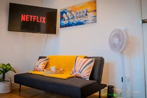 Olohuoneessa on musta sohva, keltainen heittopeitto, taulu, televisio, jossa näkyy teksti »NETFLIX», ja lattiatuuletin.