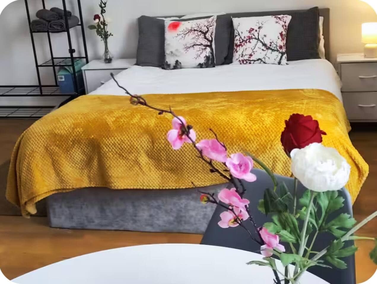 A bedroom with a bed, a bedside table, a nightstand, and a wall-mounted shelf with towels.