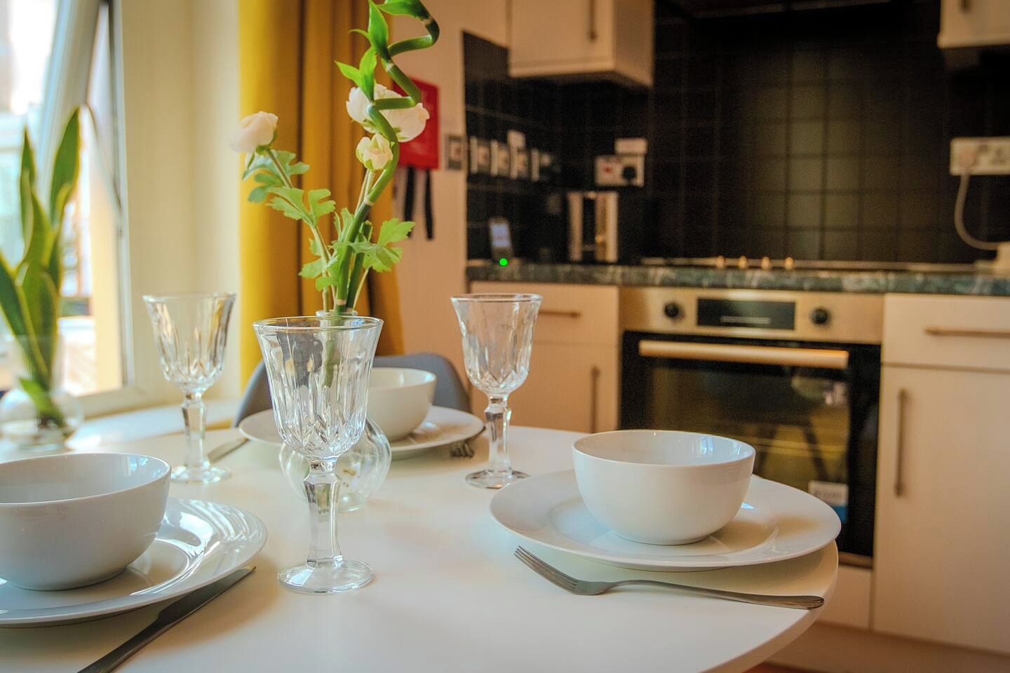 A dining table set with white plates, bowls, and glassware in a modern kitchen with a tiled backsplash and stainless steel appliances.