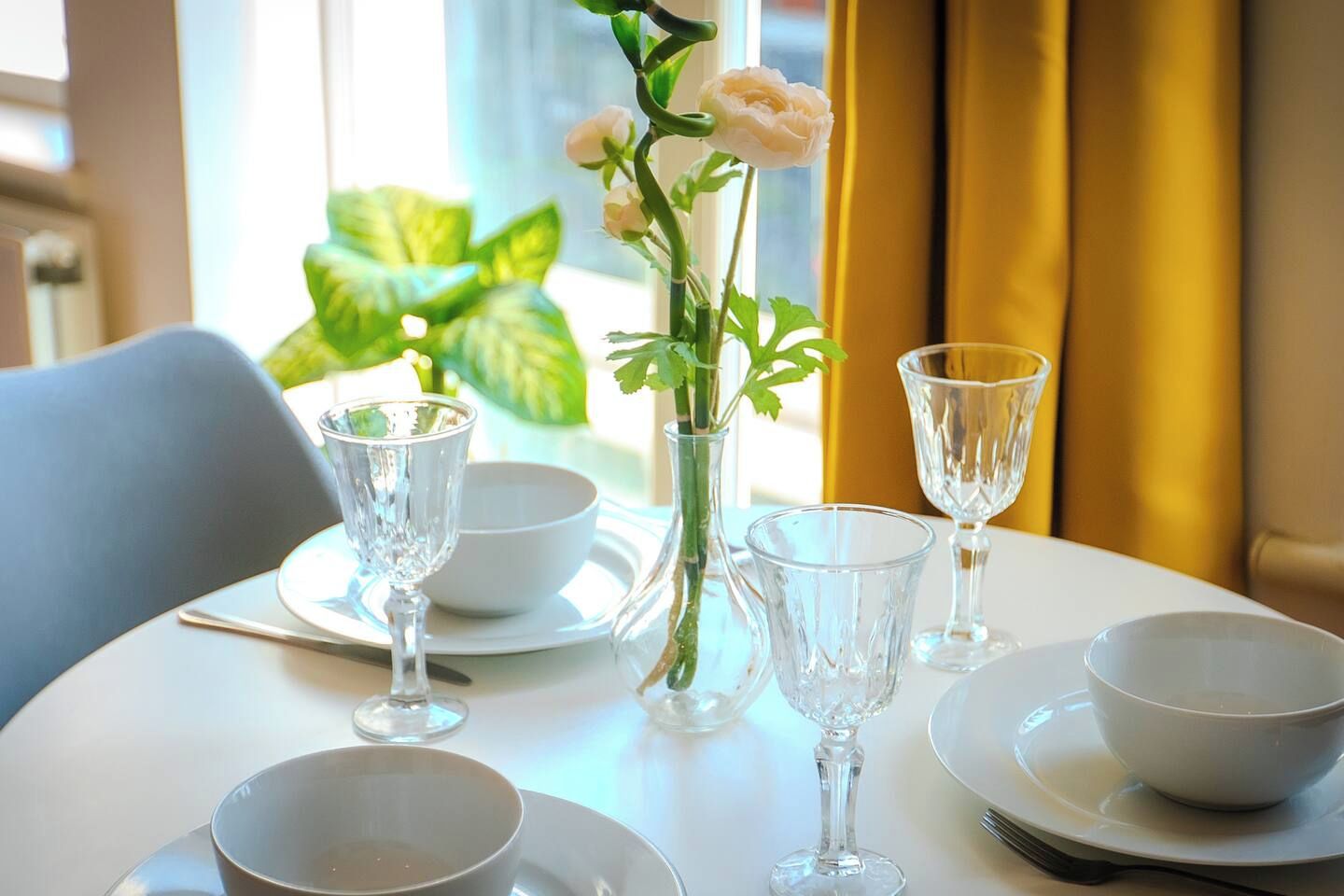 A table set for dining with clear glassware, a vase with flowers, and a view of a plant through a window.