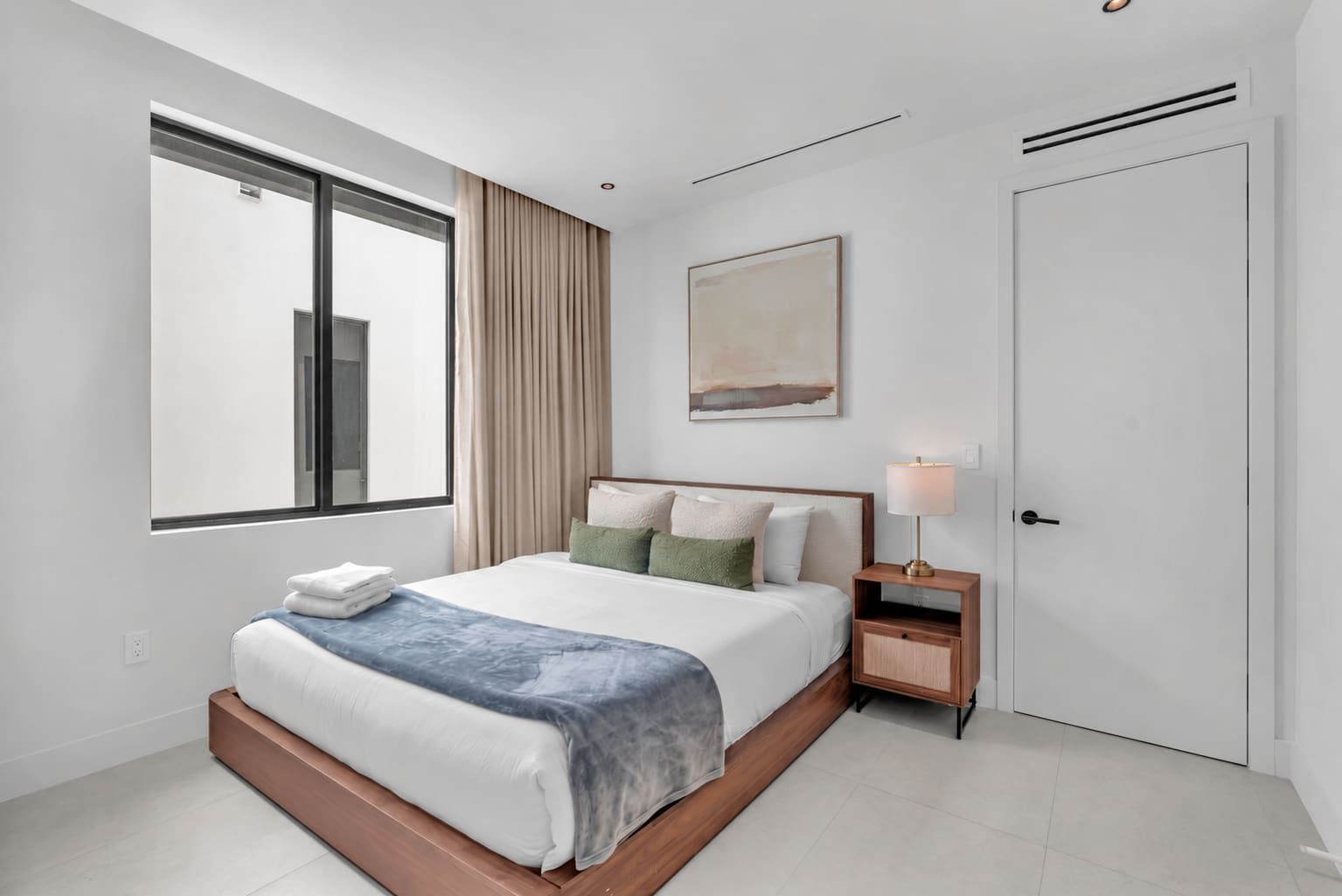 Une chambre d’hôtel moderne avec un grand lit, une table de chevet avec une lampe, une fenêtre avec des rideaux et une porte.
