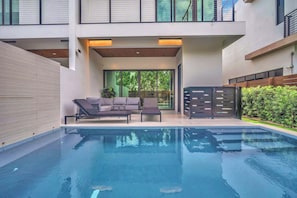 A modern outdoor pool area with a lounge chair, a sofa, and a wooden deck. - Miura New Modern Miami | Luxury Villa  4BR 10PPL (Miami)