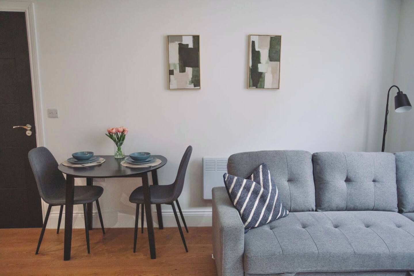 A modern living room with a grey sofa, two black chairs, a small round table with a vase of flowers, and two abstract paintings on the wall.