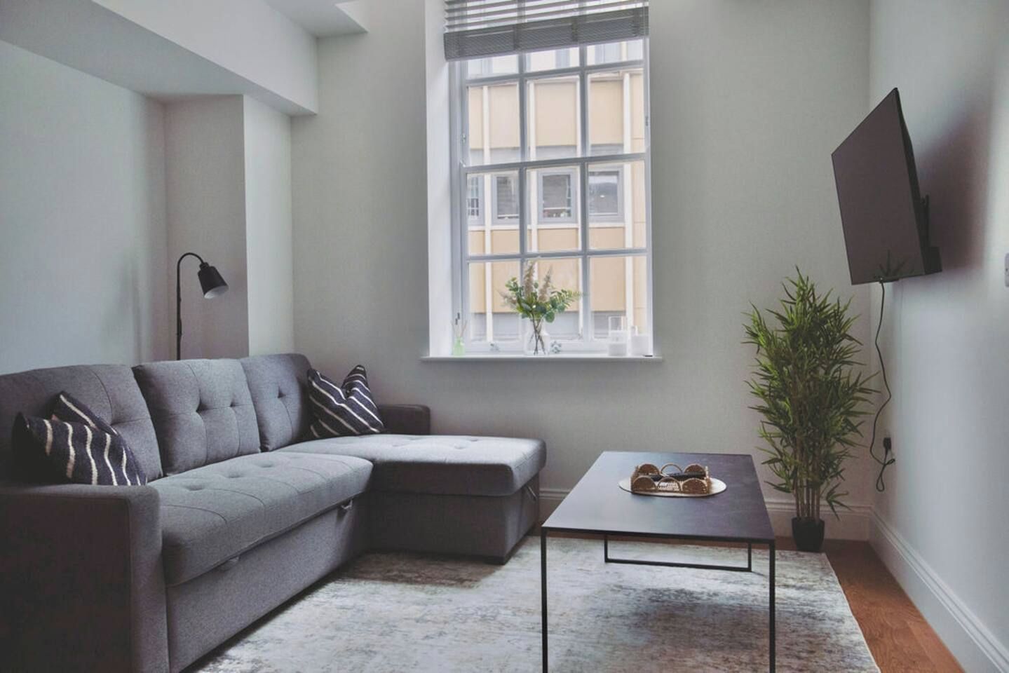 Un salon moderne avec un canapé d’angle gris, une petite table, une plante en pot et un téléviseur fixé au mur.