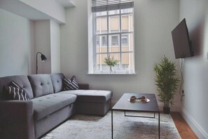 Ein modernes Wohnzimmer mit einem grauen Ecksofa, einem kleinen Tisch, einer Topfpflanze und einem Fernseher an der Wand.