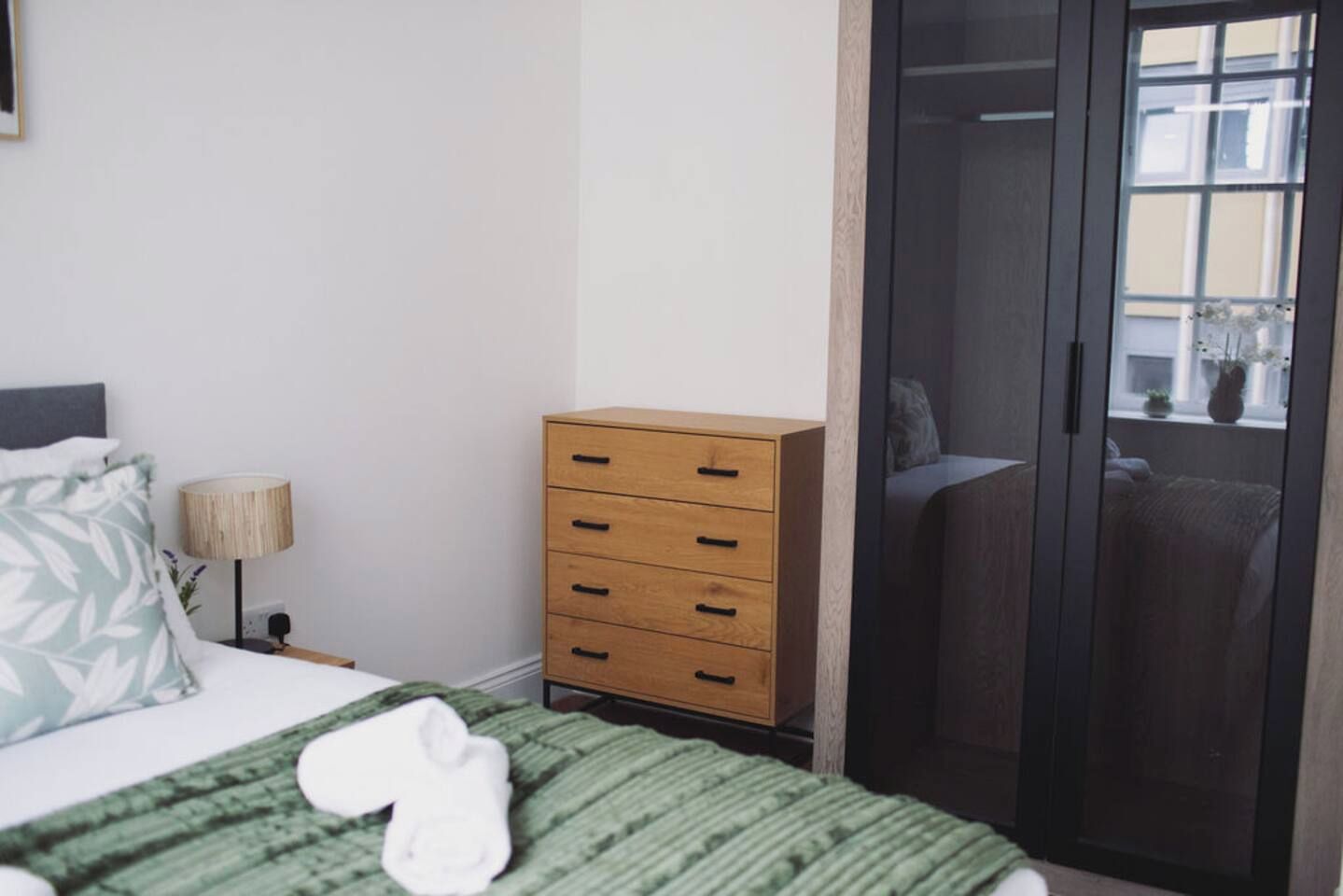 A bedroom with a bed, a wooden dresser, a nightstand, a lamp, and a wardrobe.
