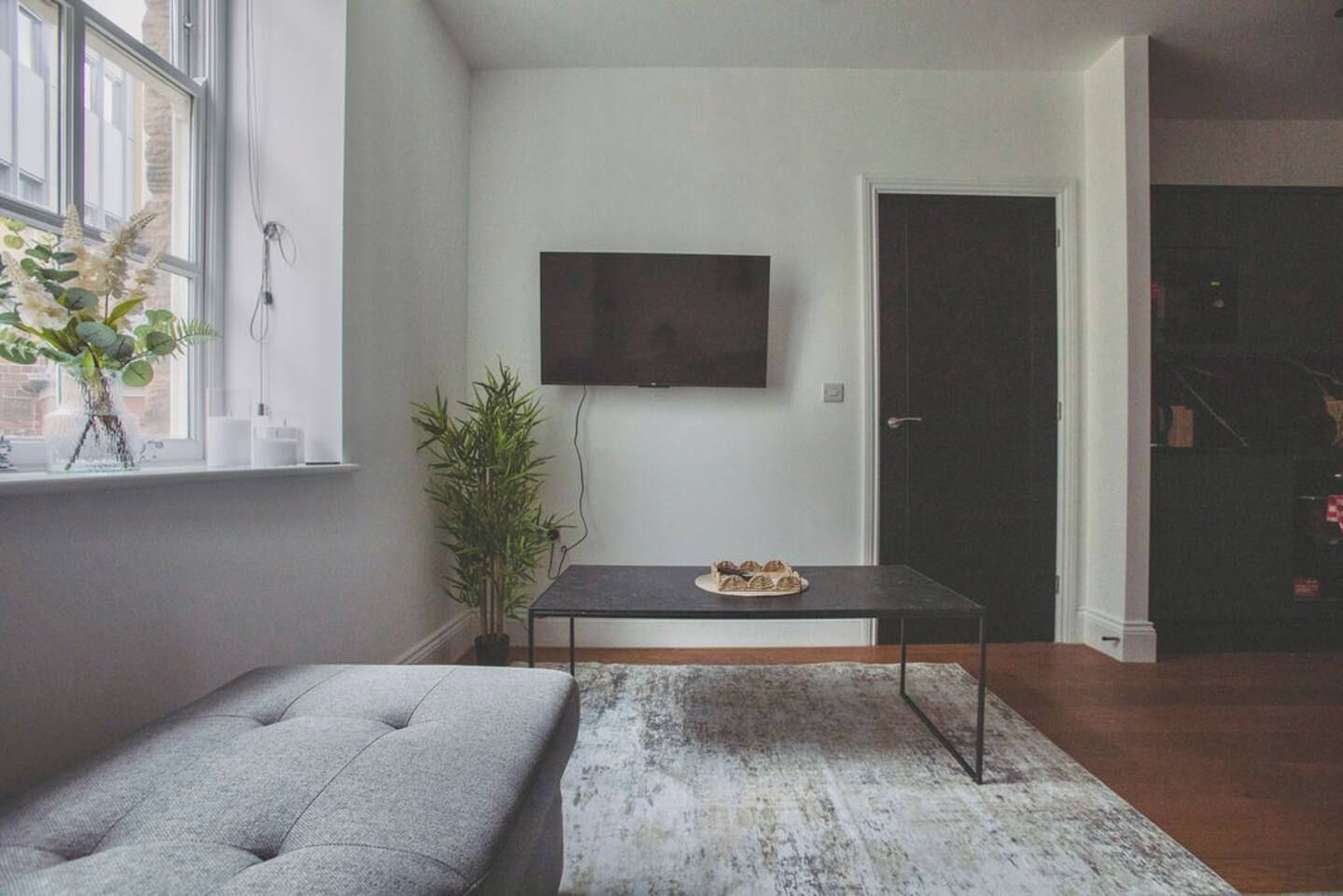 A living room with a sofa, a table with a plate of food, a potted plant, and a TV on the wall.