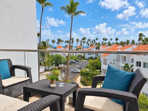 A balcony with wicker furniture, a glass railing, and a view of a street with palm trees and buildings. - Ocean View Sunrise 2BR Navio Los Corales C403 (Bávaro)