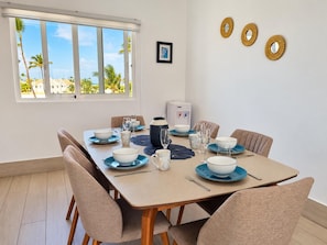 A dining area with a table set for four, a water dispenser, and a view of palm trees through a window. - Ocean View Sunrise 2BR Navio Los Corales C403 (Bávaro)