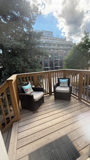 Een houten terras met twee rieten stoelen en kussens, met uitzicht op een gebouw dat bepalen is.