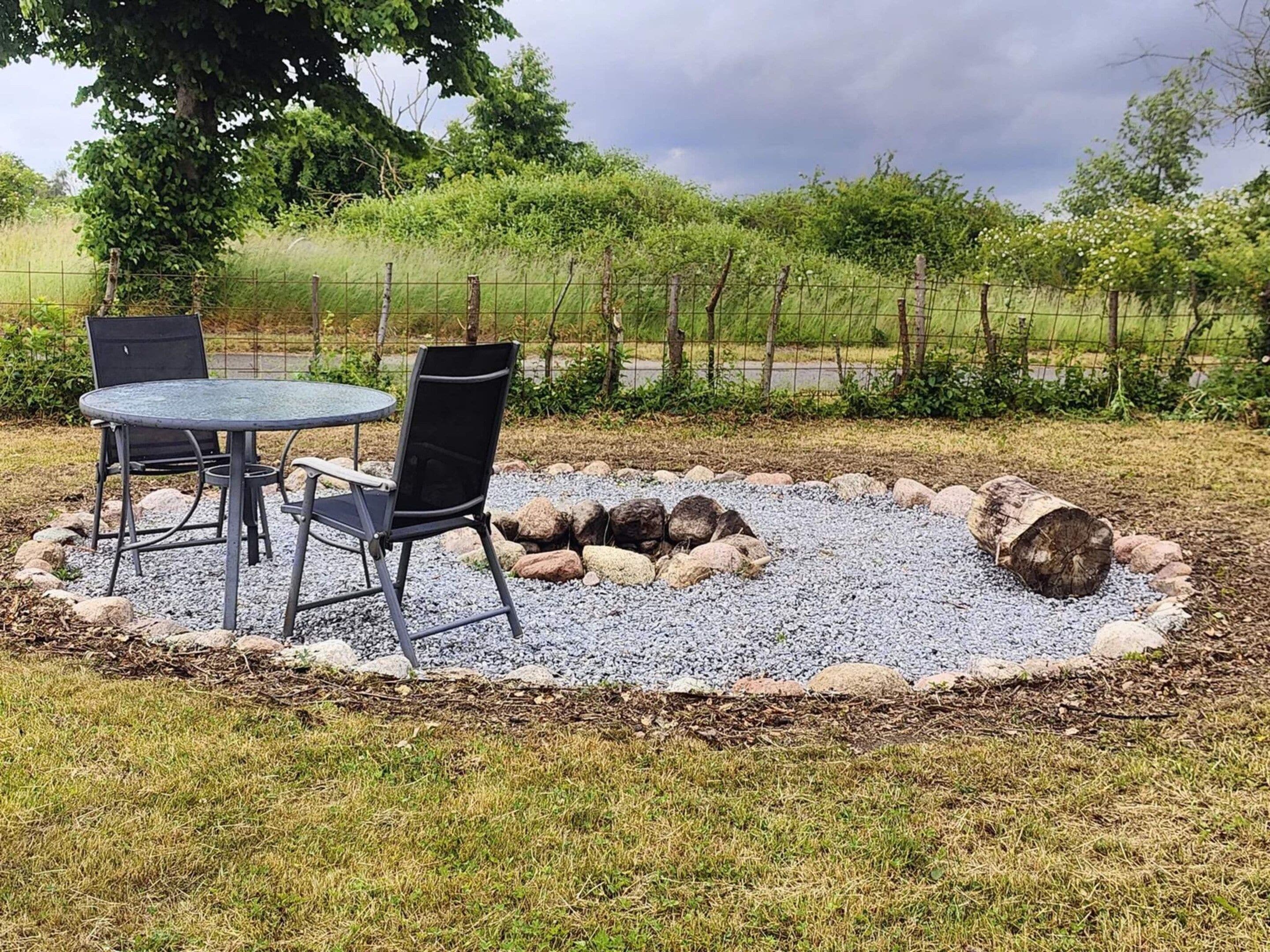 Een terras met een ronde tafel en twee stoelen, omgeven door een grindpad met stenen en boomstammen.