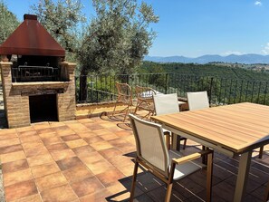 Een terras buiten met een bakstenen barbecue, rieten stoelen en een houten tafel.