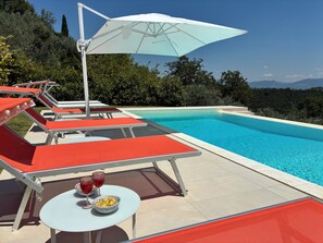 A poolside area with lounge chairs, a table with a bowl of chips and two glasses of red wine, and a large umbrella providing shade. - Stunning villa with a pool in the Umbria Valley (Gualdo Cattaneo)