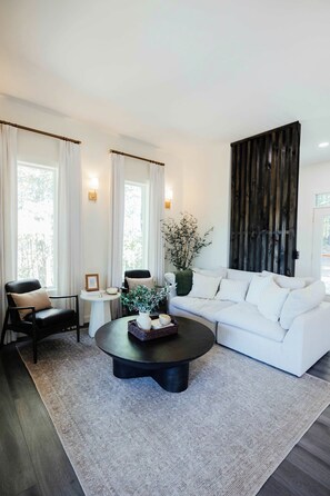 A modern living room with a white sofa, a black coffee table, and a large window with curtains. - Dark Skies | Rooftop Deck | Hot Tub | Pond Views | Game Room! (Campton)