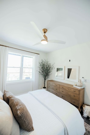Een slaapkamer met een bed, een dressoir, een plafondventilator, een raam en een plant.
