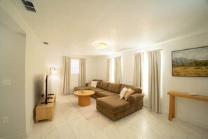Un salon comprenant un canapé d’angle, une table basse ronde, un téléviseur posé sur un meuble, un tableau accroché au mur et une console en bois.