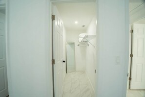 Un couloir aux murs blancs, doté d’un sol en marbre et d’une porte donnant sur une pièce où se trouvent un miroir et une étagère blanche.