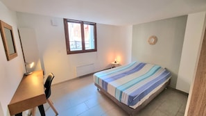 A bedroom with a bed, a desk, a chair, a window, and a wall-mounted light fixture. - Le sétois 2, intimiste et cosy (Sète)