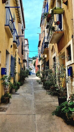 A narrow street flanked by buildings with balconies, potted plants, and outdoor seating areas. - Le sétois 2, intimiste et cosy (Sète)