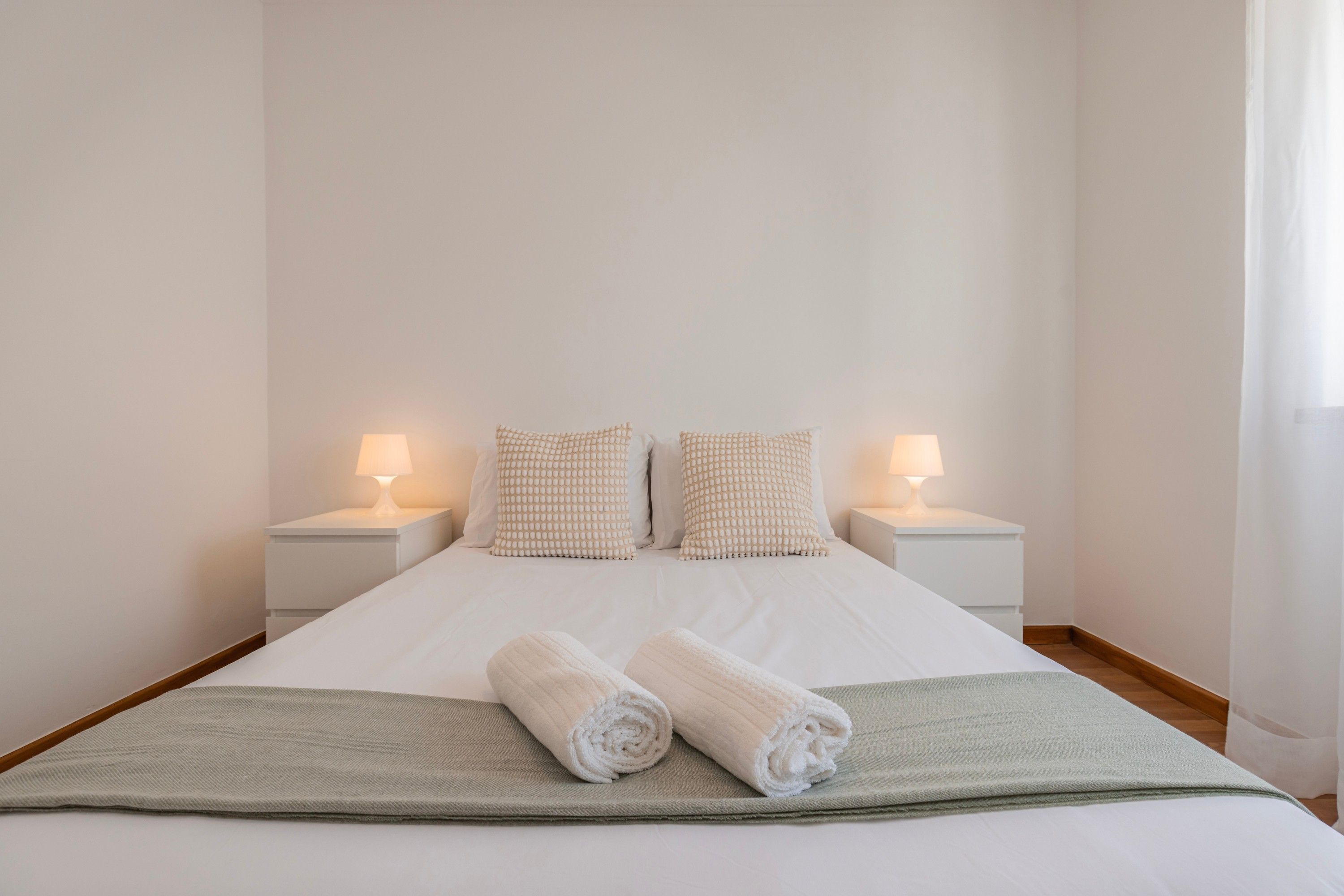 A neatly made bed with two pillows and two rolled towels, flanked by bedside tables with lamps.