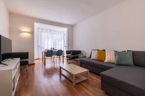 A modern living room with a flat-screen TV, a sofa with patterned cushions, a wooden coffee table, and a dining area with chairs and a table. - Apartment 'Lisbon Oceanside' (Carcavelos)