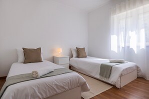 Habitación de hotel con dos camas individuales, una mesita de noche con lámpara, ventana con cortinas translúcidas y piso de madera.
