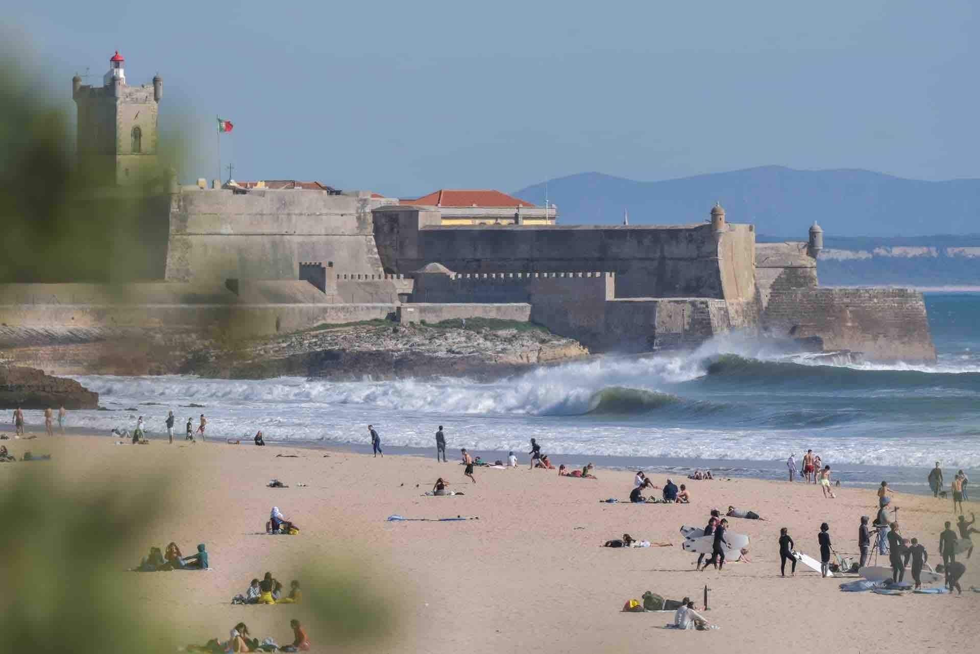 A beach with waves crashing onto the shore, a castle-like structure in the background, and people on the sand.