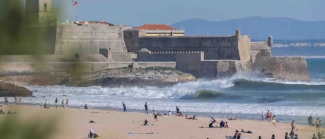 A beach with waves crashing onto the shore, a castle-like structure in the background, and people on the sand.