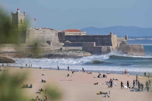 A beach with waves crashing onto the shore, a castle-like structure in the background, and people on the sand. - Apartment 'Lisbon Oceanside' (Carcavelos)