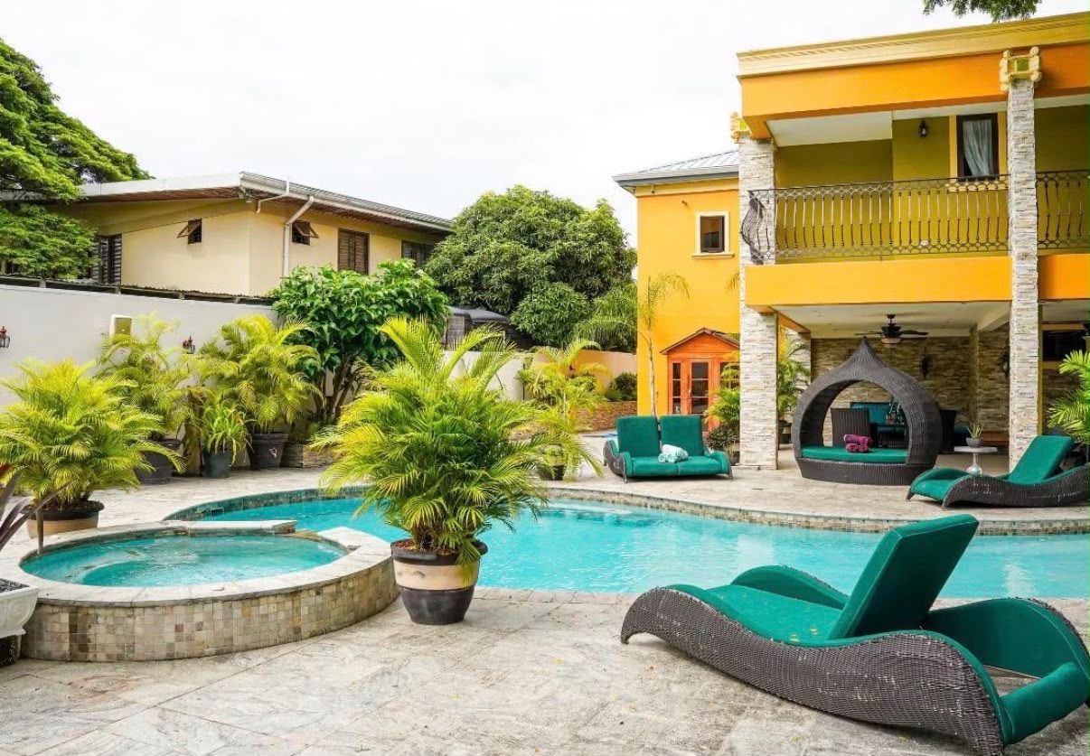Une piscine avec des chaises longues, des plantes en pot et un bâtiment doté d’un balcon.