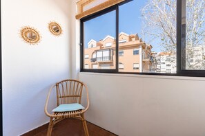 Ein Zimmerwinkel mit einem Korbstuhl, einem Fenster mit Blick auf Gebäude und zwei Wanddekorationen.