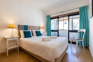 A hotel room with a large bed, two bedside tables with lamps, a chair, and a view of buildings through the window. - Apartment 'Cozy Carcavelos Apartment' (Carcavelos)