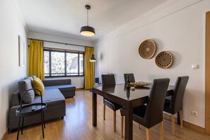 Ein modernes Wohnzimmer mit einer Couch, einem Esstisch mit Stühlen, einem Fernseher und einem Fenster mit Vorhängen.