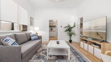 Ein modernes Wohnzimmer mit einem grauen Sofa, einem weißen Couchtisch, einem gemusterten Teppich, einem Fernseher und einer Topfpflanze.