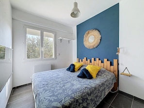 A bedroom with a bed, a window, a wall-mounted mirror, and a light fixture. - Le Cormoran, cosy vue mer. (Frontignan)