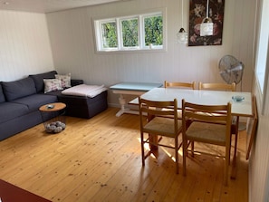 A living room with a sofa, a small round table, a dining table with chairs, a window with a view of greenery, and a wall-mounted fan. - 4 Star Holiday Home in Allinge (Allinge)