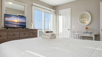 Une chambre avec un grand lit, une commode, une télévision, une fenêtre donnant sur un beau paysage, une porte et une petite table sur laquelle est posé un vase.