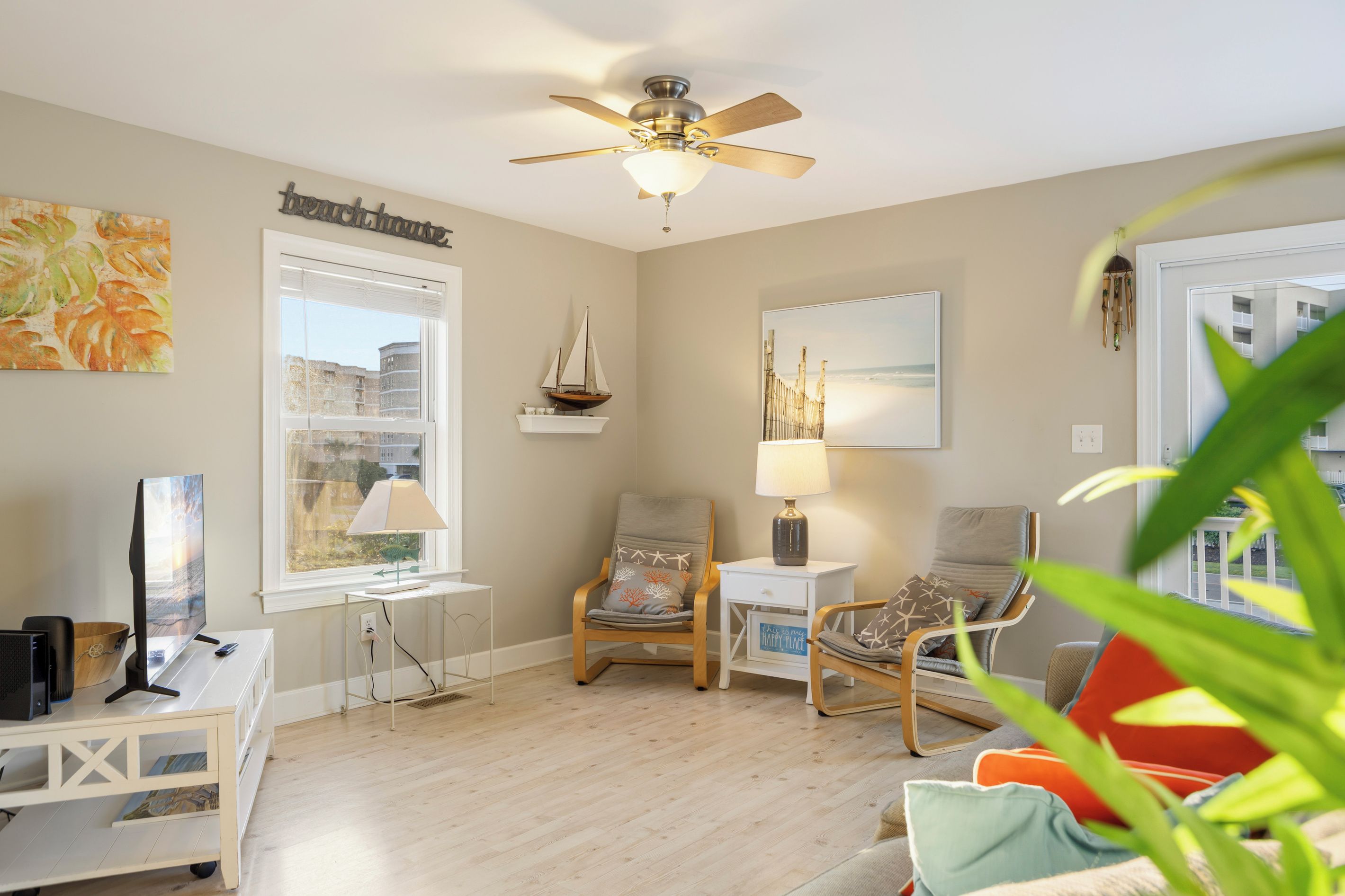 A living room with a ceiling fan, a sailboat decoration, a beach-themed painting, and a view of buildings through a window.
