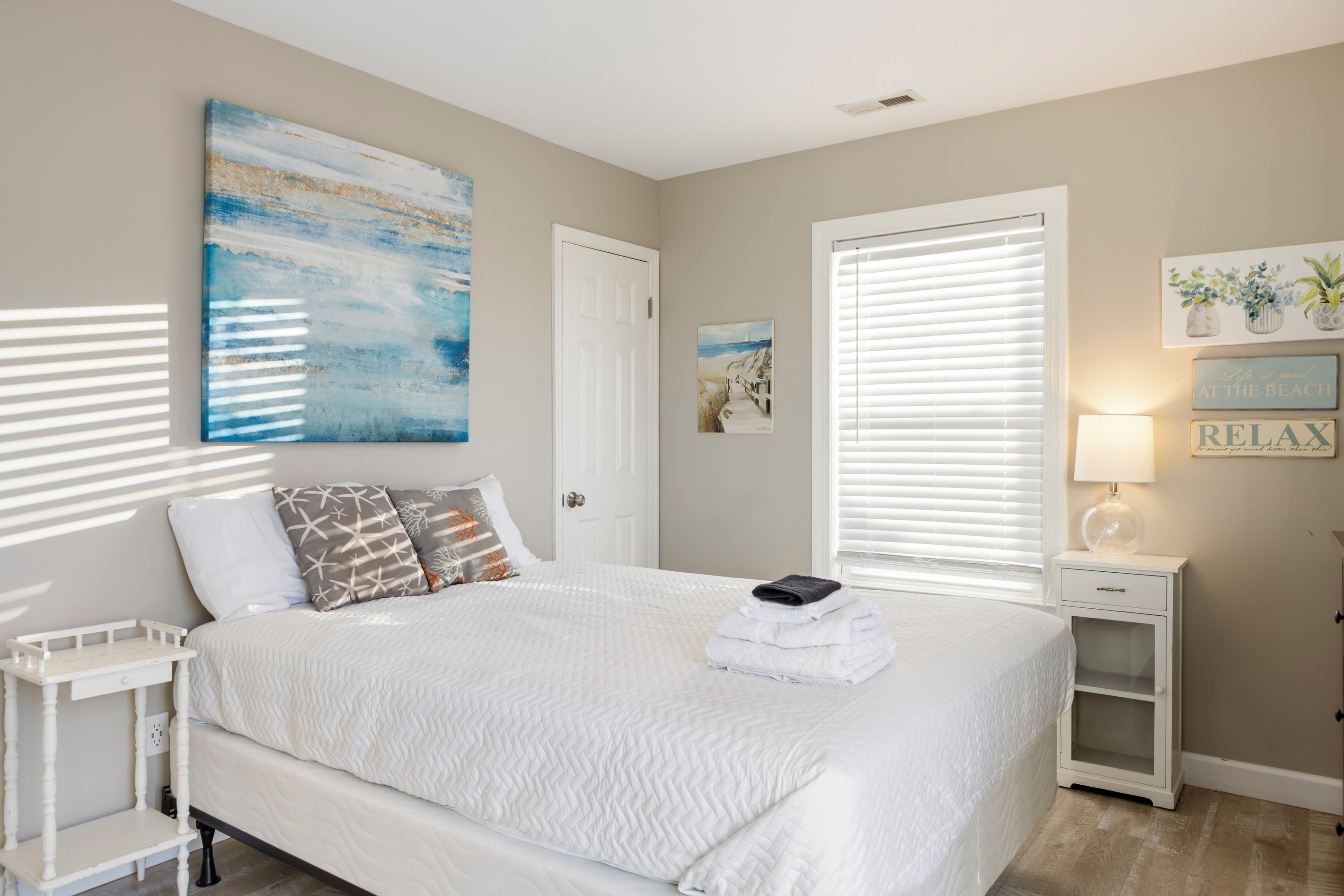 A bedroom with a bed, a bedside table, a nightstand, a window with blinds, and wall art.